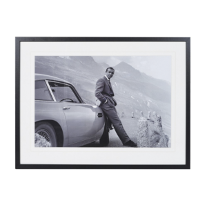 The iconic classic picture featuring Sean Connery as 007 James Bond leaning against his Aston Martin Car in the Scottish Highlands setting. Black & white photo in black framing and passe partout Measurements: Dimensions: H:63 W:83 cm.